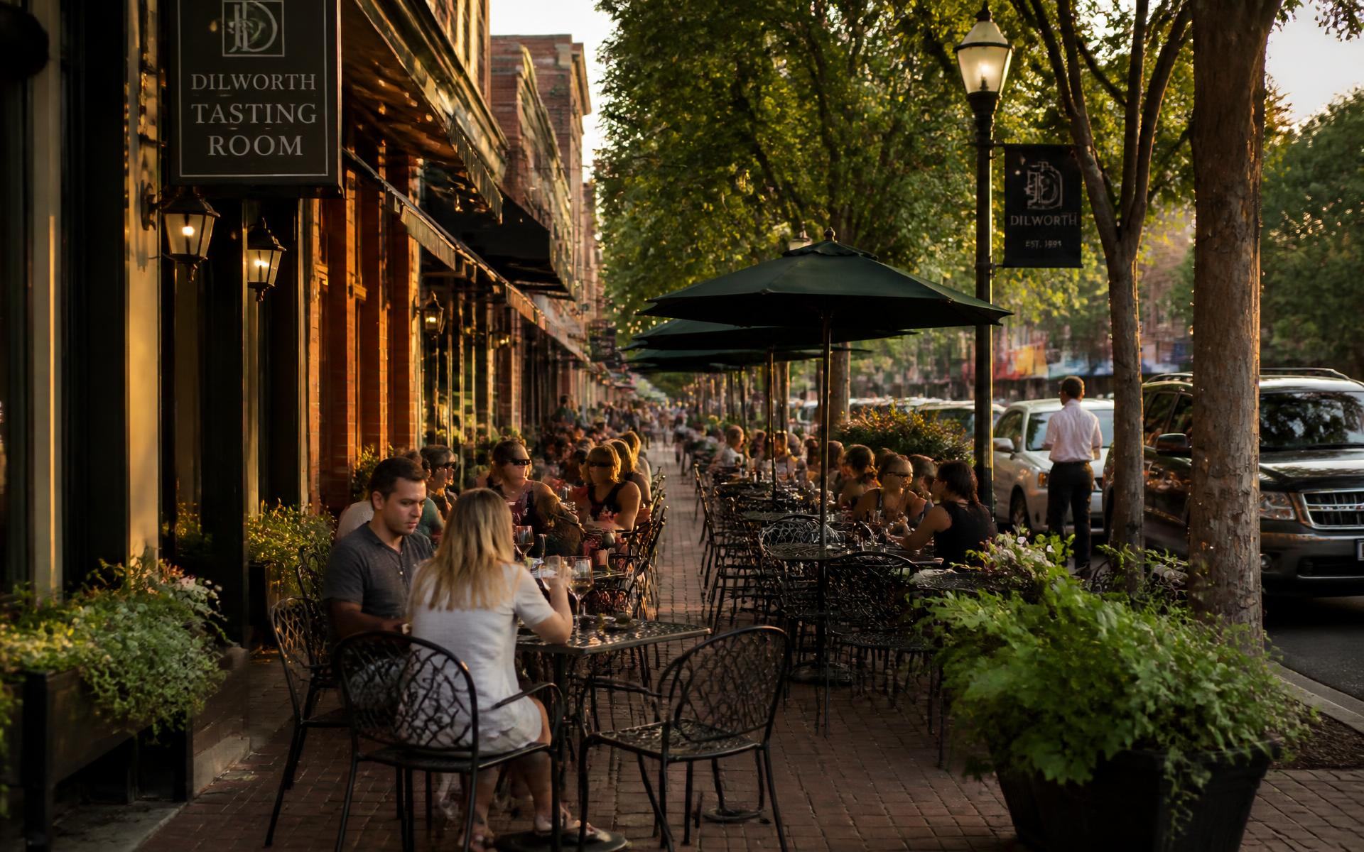 Diners at wrought-iron patio tables under string lights on a tree-shaded sidewalk in Dilworth, Charlotte